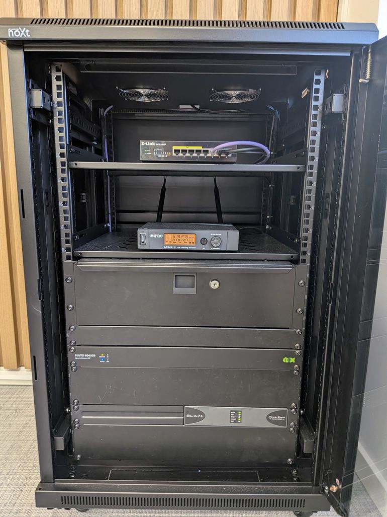 Tall black 4‑post server rack with equipment mounted: a D‑Link network switch on a middle shelf, a compact gray MIPRO/UPS device on the next shelf, and a large Blaze power unit at the bottom inside a cabinet.