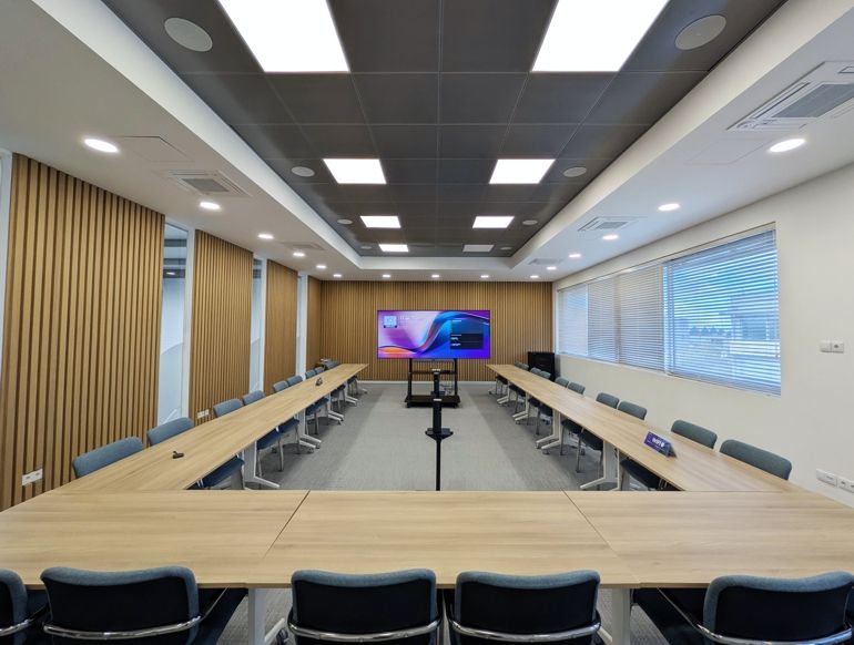 Empty modern conference room with long wooden tables arranged in a U-shape and a large screen on a stand at the front.