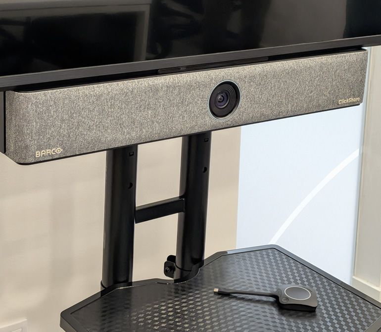Barco ClickShare video collaboration camera bar mounted on a stand above a textured black table, with a small black remote-like device on the table in front.