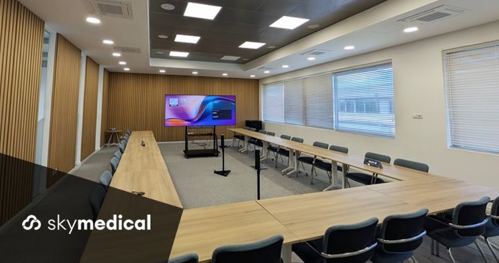 Modern conference room with U-shaped wooden tables, grey chairs, and a large screen displaying a colorful slide; window blinds along the right wall.