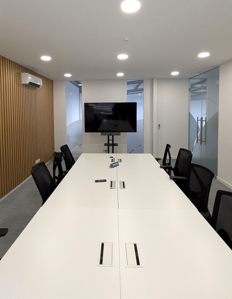 Modern conference room with a long white table, black chairs, and a mounted TV screen at the far end.
