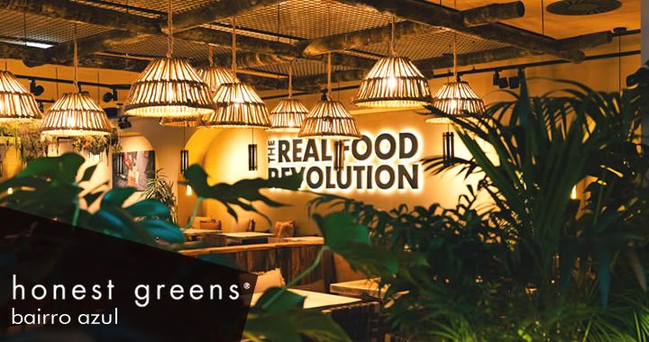 Interior of Honest Greens, Barrio Azul, with hanging wicker pendant lights and lush plants; wall reads 'The Real Food Revolution'.