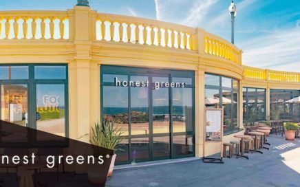 Exterior view of a bright yellow Honest Greens storefront with glass windows and outdoor seating under umbrellas on a sunny day