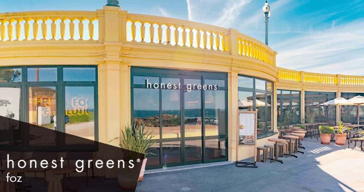 Exterior view of a bright yellow Honest Greens storefront with glass windows and outdoor seating under umbrellas on a sunny day