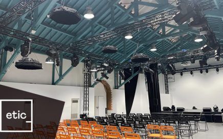 Event venue set up for a talk: orange chairs facing a stage with black curtains and a long desk, under a teal ceiling with rigging and lights, logo 'etic' in corner.
