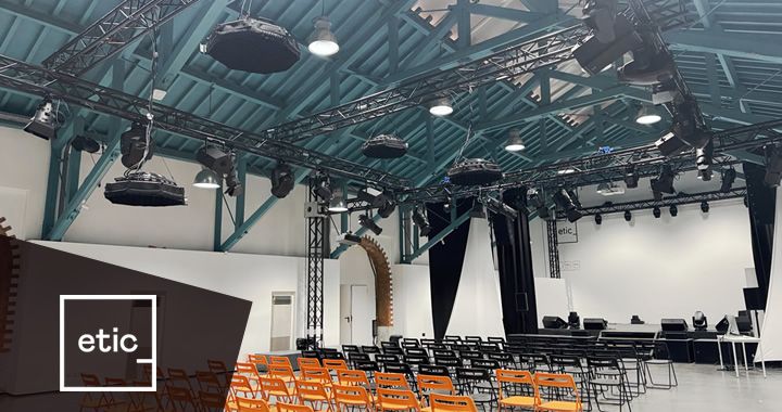 Event venue set up for a talk: orange chairs facing a stage with black curtains and a long desk, under a teal ceiling with rigging and lights, logo 'etic' in corner.