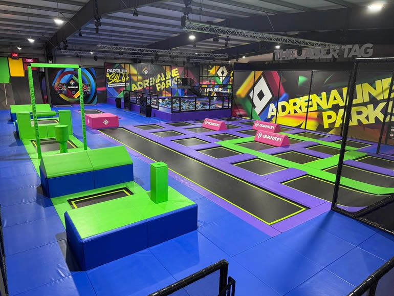 Indoor trampoline park with purple and green trampolines, padded platforms, and netted enclosures; colorful wall mural says Adrenaline Park.