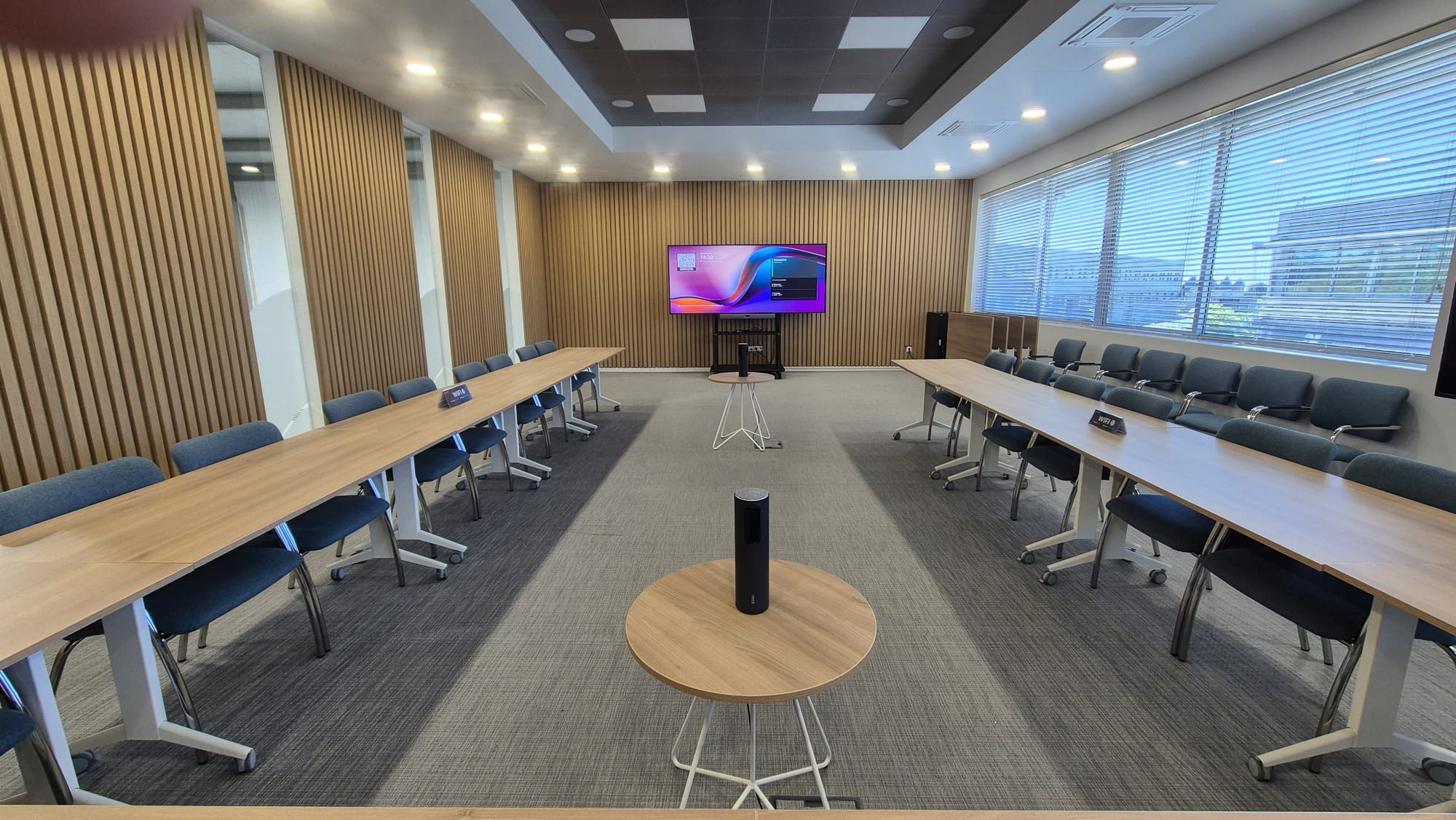 Conference room set for a meeting with long U-shaped wooden tables and blue chairs, a central screen on a wood-paneled wall, and large windows with blinds to the right.
