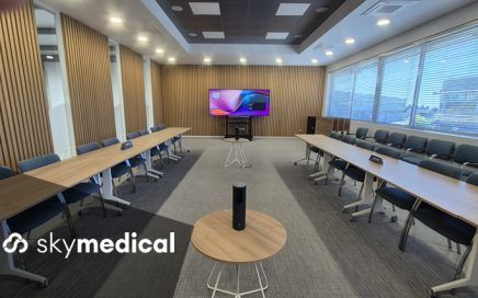 Empty modern conference room arranged in a U-shaped table setup with blue chairs, a large wall screen at the far end, and a small round table holding a speaker in the foreground; skylmedical branding visible in the bottom-left corner.