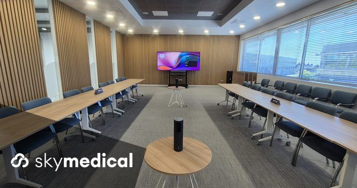 Empty modern conference room arranged in a U-shaped table setup with blue chairs, a large wall screen at the far end, and a small round table holding a speaker in the foreground; skylmedical branding visible in the bottom-left corner.