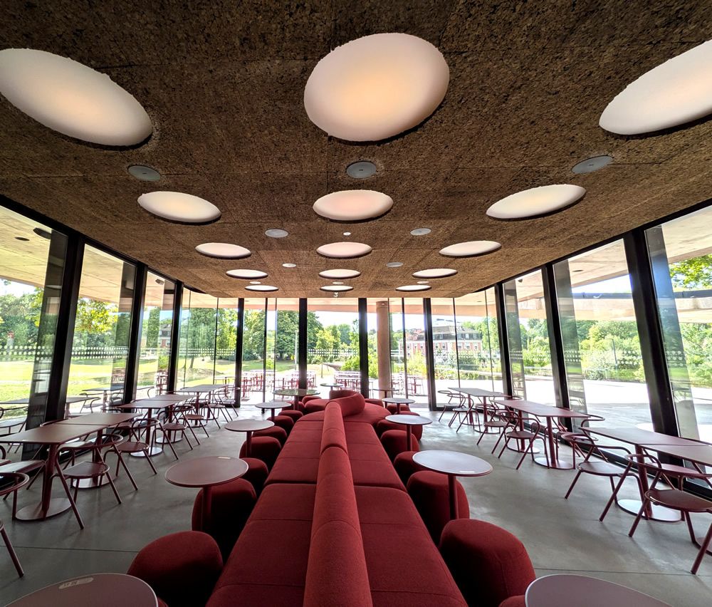 Long red cushioned seating area with round white tables in a glass-walled cafe overlooking a green lawn.