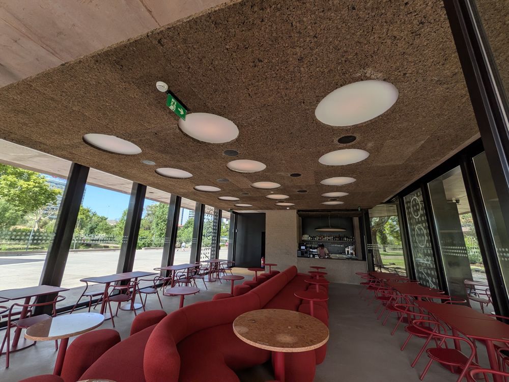 Modern cafe interior with red curved couches, round wooden tables, and red chairs along large glass walls overlooking a sunny outdoor area.