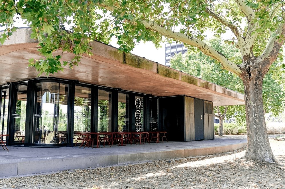 Café with glass walls and red metal tables under a tree, outdoor seating along a curved concrete platform.