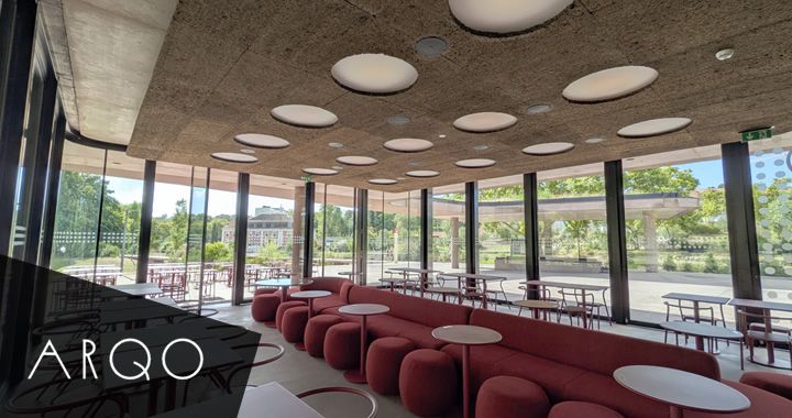 Modern glass-walled lounge with red ottoman seating and small white tables, circular ceiling lights overhead. ARQO branding visible in the corner.