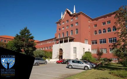 Exterior view of a red-brick school building with a white entrance stair and parked cars in a lot under a clear blue sky; blue school logo in bottom-left corner.