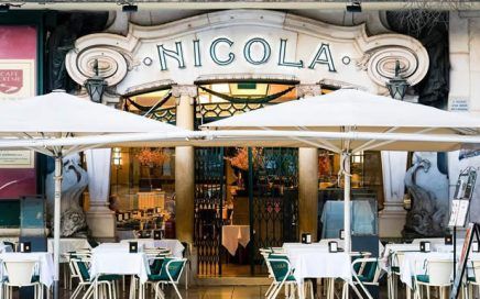 Front of a restaurant named NICOLA with outdoor seating under white umbrellas and white-clothed tables, inviting entry doors in the background.