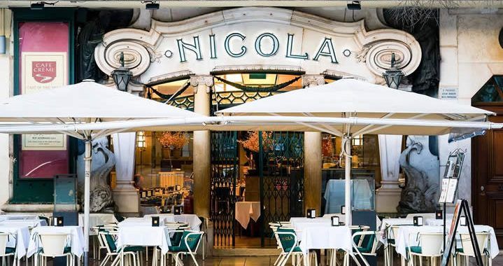 Front of a restaurant named NICOLA with outdoor seating under white umbrellas and white-clothed tables, inviting entry doors in the background.
