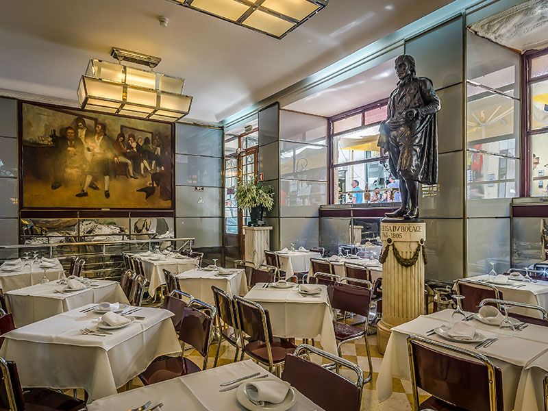 Upscale restaurant interior with white-tablecloth tables, brown chairs, and a bronze statue on a pedestal nearby.